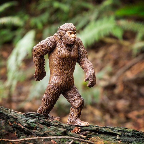 A Bigfoot action figure walking on a log in a forest setting, surrounded by greenery.