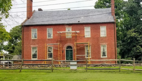 Historic red brick house with scaffolding in front, surrounded by green grass and a wooden fence.