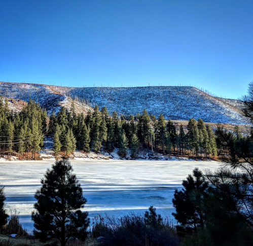 A serene winter landscape featuring a frozen lake surrounded by snow-covered hills and evergreen trees under a clear blue sky.