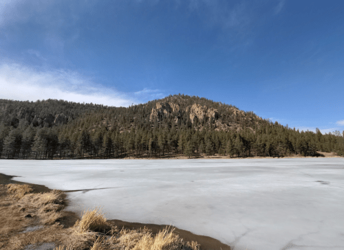 A frozen lake surrounded by pine trees and a rocky hillside under a clear blue sky.