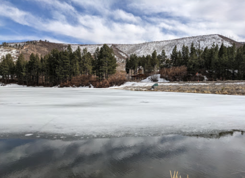 A frozen lake surrounded by snow-covered hills and evergreen trees under a partly cloudy sky.