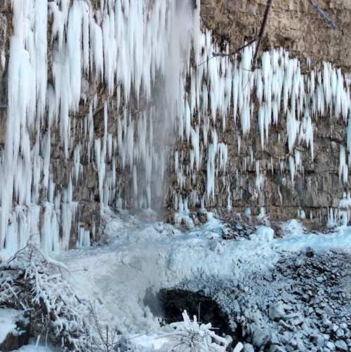 Icicles hang from a rocky cliff, with a frozen waterfall and icy ground below, creating a winter landscape.