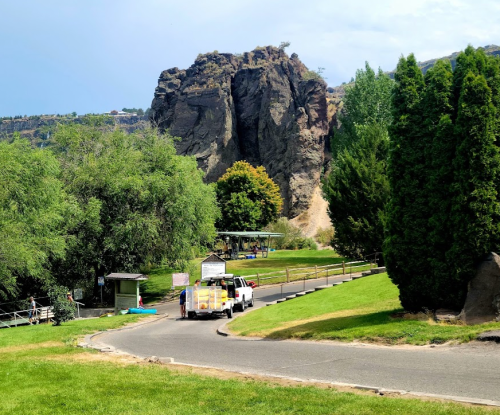 A truck drives along a winding road near a rocky cliff, surrounded by greenery and trees under a blue sky.