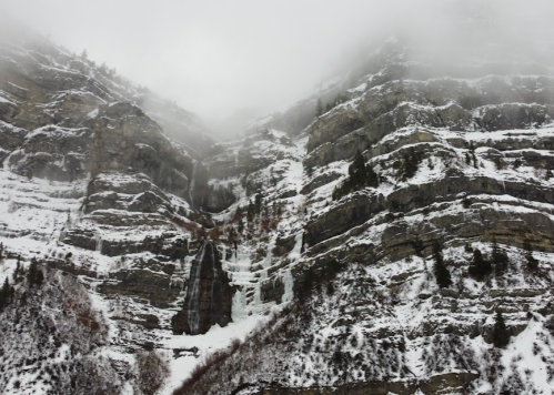 Snow-covered mountain cliffs shrouded in mist, with a waterfall cascading down the rocky face.