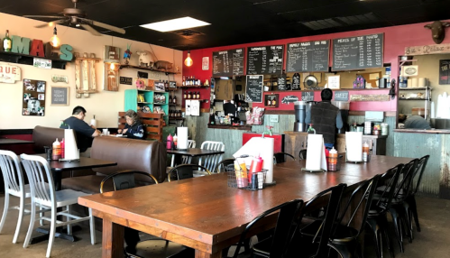 A cozy restaurant interior with wooden tables, black chairs, and a menu board, featuring customers dining and staff at the counter.