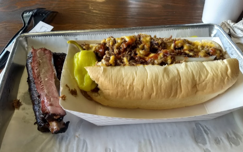 A tray with a loaded hot dog topped with beef and cheese, alongside a slice of smoked brisket.