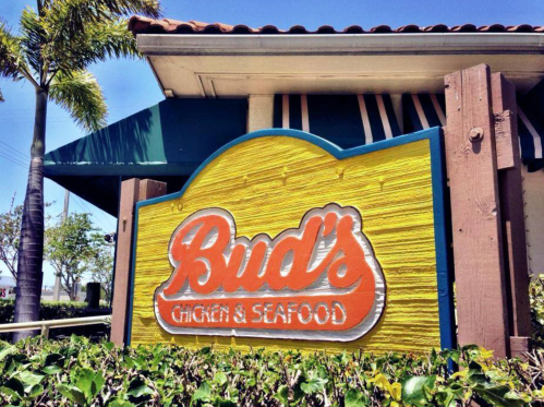 Sign for Bud's Chicken & Seafood, featuring a colorful design, surrounded by greenery and palm trees.