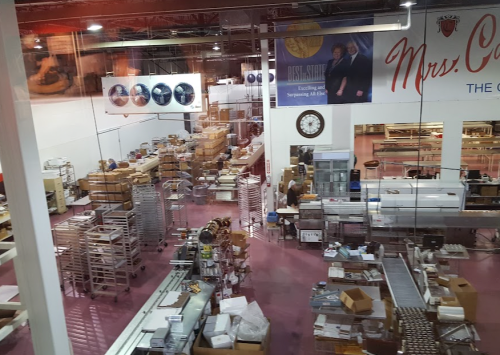 A large industrial kitchen filled with shelves, baking equipment, and packaging materials, with a pink floor and banners.