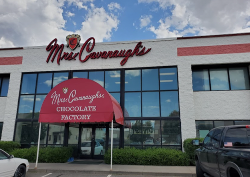 Exterior of Mrs. Cavanaugh's Chocolate Factory, featuring a red awning and large windows under a cloudy sky.