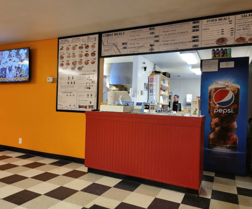 Interior of a casual eatery with a red counter, menu on the wall, and a TV showing images. Pepsi fridge on the side.