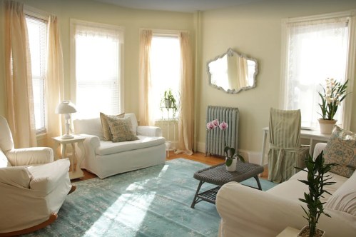 Bright living room with light yellow walls, white furniture, a blue rug, and large windows letting in natural light.