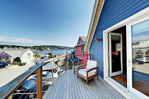 A sunny balcony with a chair overlooks a harbor and colorful houses under a clear blue sky.