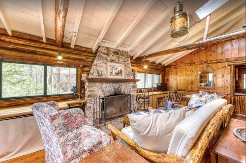 Cozy log cabin interior with a stone fireplace, wooden beams, and comfortable seating. Natural light from large windows.