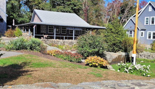 A quaint house named "Paper House" surrounded by greenery and colorful flowers on a sunny day.