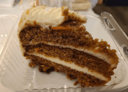 A slice of carrot cake with cream cheese frosting, displayed in a clear plastic container.