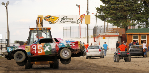 A tow truck lifts a colorful, damaged car at Sycamore Speedway, with spectators and other vehicles in the background.