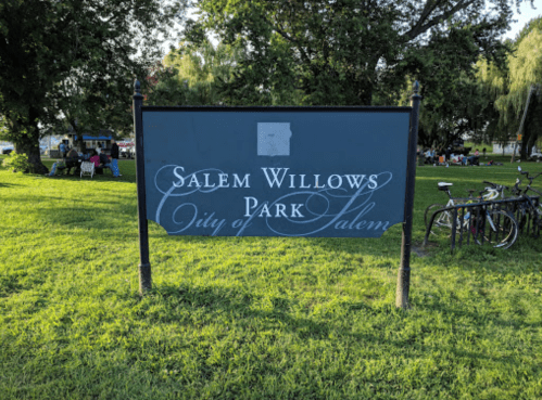 Sign for Salem Willows Park in Salem, surrounded by green grass and trees, with people and bicycles in the background.