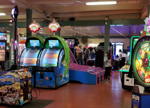 A colorful arcade with various games, including a slot machine and a spinning wheel, and people playing in the background.