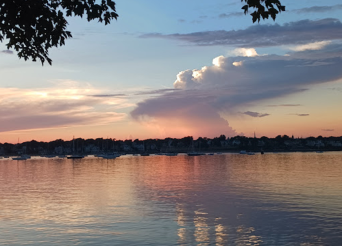 A serene sunset over a calm water body, with colorful clouds reflecting on the surface and silhouettes of boats in the distance.