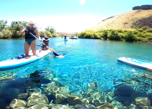 Two people paddleboarding in clear blue water, surrounded by greenery and a rocky hillside under a bright sun.