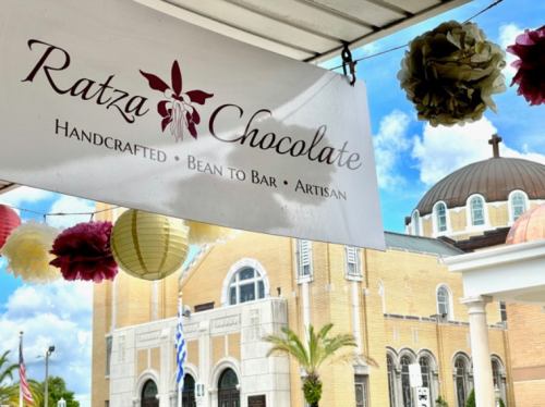 Sign for Ratza Chocolate, featuring "Handcrafted Bean to Bar Artisan," with a church and decorative lanterns in the background.