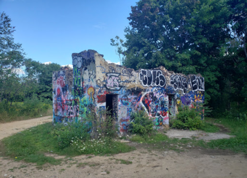 A small, graffiti-covered ruin surrounded by greenery and a dirt path.