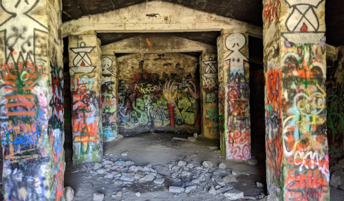 Graffiti-covered walls and pillars inside a dilapidated building, with debris scattered on the ground.