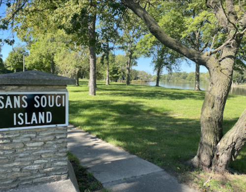 Sign for Sans Souci Island beside a pathway, with lush green trees and a river in the background.