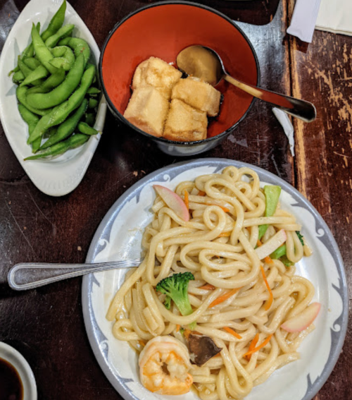 A plate of stir-fried noodles with shrimp and vegetables, accompanied by edamame and a bowl of tofu in sauce.
