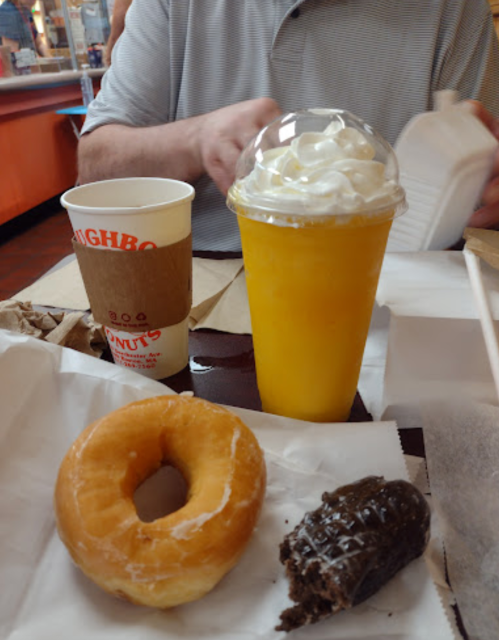 A table with a glazed donut, a chocolate donut, a cup of orange drink topped with whipped cream, and a coffee cup.