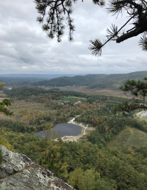 A scenic view from a rocky overlook, showcasing a valley with trees and a pond under a cloudy sky.