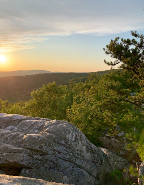 A scenic view of a sunset over a lush green landscape, with rocky foreground and trees silhouetted against the sky.