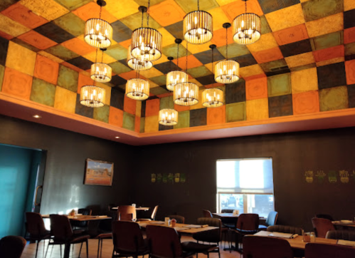 A cozy restaurant interior featuring a colorful patterned ceiling and stylish pendant lights, with empty tables and chairs.