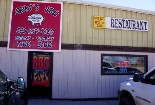 Exterior of Greg's BBQ restaurant with a sign displaying hours and contact information. BBQ-themed door design visible.