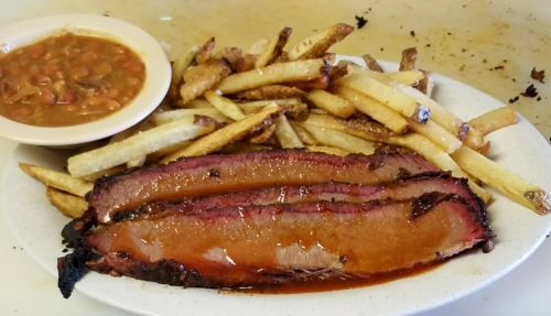 A plate of sliced brisket with barbecue sauce, served with French fries and a side of baked beans.