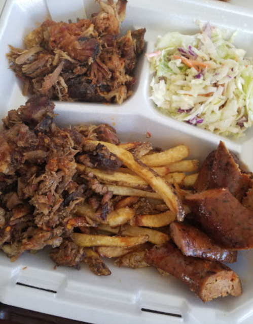 A takeout container with pulled pork, sausage, fries, and coleslaw.