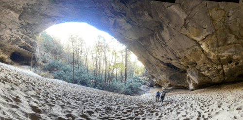 Two hikers explore a sandy cave with a large arch, surrounded by trees and sunlight filtering through.