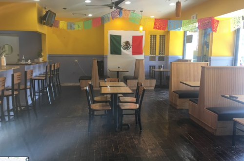 Interior of a restaurant with yellow walls, wooden tables, and a Mexican flag displayed. Colorful banners hang from the ceiling.