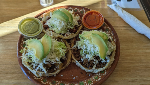 A plate of four tacos topped with avocado, lettuce, cheese, and served with two sauces on the side.