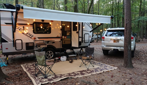 A camper with an awning and string lights, parked in a wooded area, with chairs and a rug set up outside.