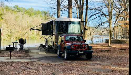 An RV parked beside a river, with a red Jeep in front, surrounded by trees and fallen leaves.