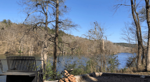 A serene river scene with bare trees and a clear blue sky, featuring a bench and a stone area by the water's edge.