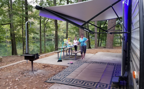 A campsite with a picnic table, two children, and a grill, surrounded by trees and a view of a lake.