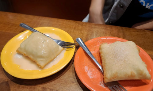 Two plates with stuffed pastries: one drizzled with sauce and the other sprinkled with sugar, both with forks beside them.
