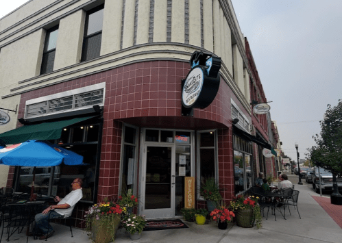 A cozy restaurant exterior with outdoor seating, colorful flowers, and an "Open" sign on a busy street.