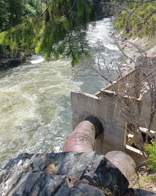 A concrete structure with a large pipe extends over a flowing river, surrounded by trees and rocky terrain.