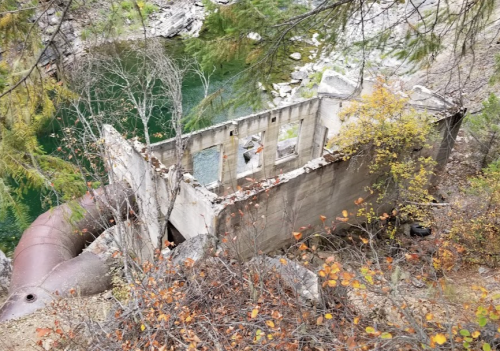 Abandoned concrete structure near a river, surrounded by trees and autumn foliage.