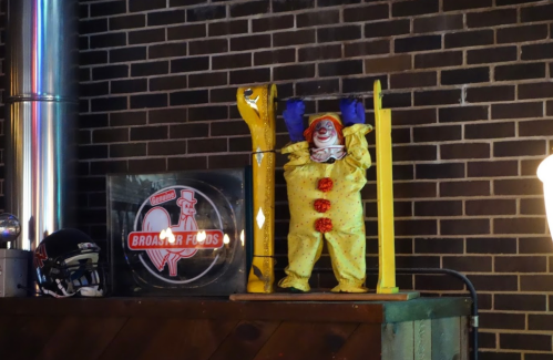 A clown figure in a yellow outfit stands in a display, surrounded by a brick wall and a sign for Broaster Foods.
