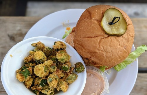 A plate featuring a sandwich with lettuce and a pickle, alongside a bowl of fried okra and dipping sauce.