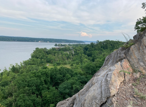 A rocky cliff overlooks a lush green landscape and a river, with distant hills under a cloudy sky.